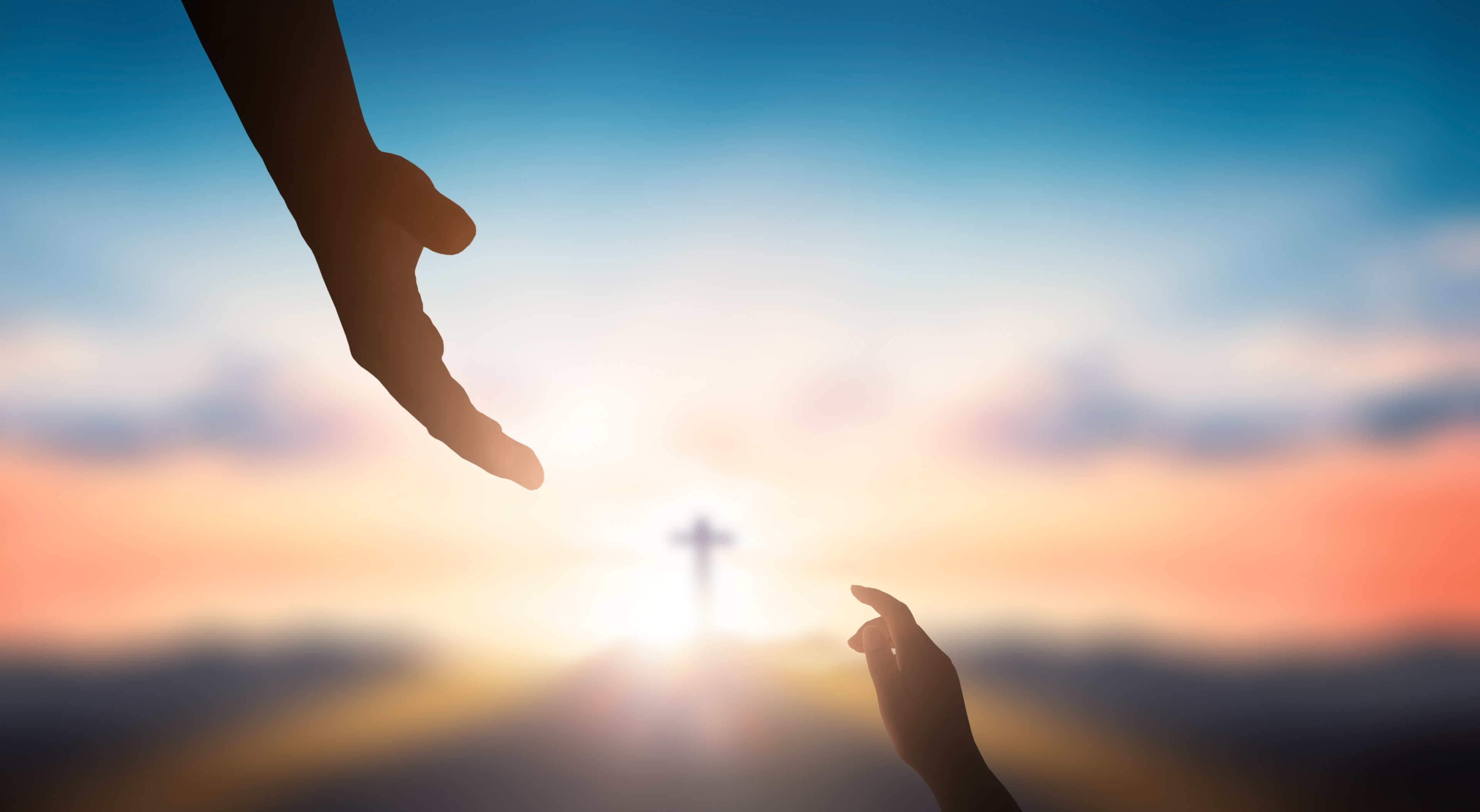 Two hands reaching toward a cross at sunset